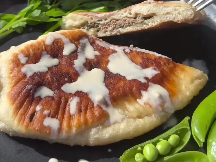 A fried cheburek drizzled with tahini on a dark plate with snap peas