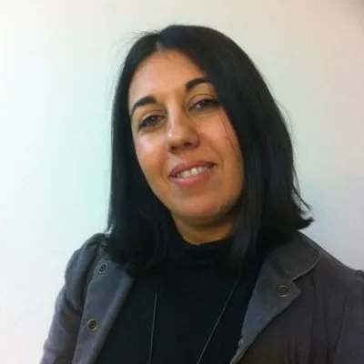 Ilanit smiling at the camera, with shoulder-length dark hair, wearing a black top and a grey jacket